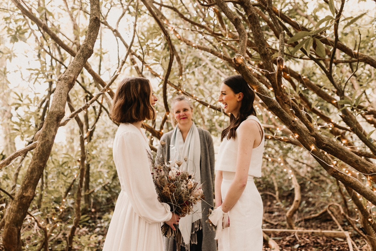 two women in wedding dresses in the woods with a celebrant 