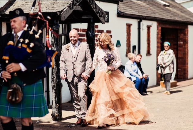 groom in check suit, bride in tiered tan skirt bagpiper playing