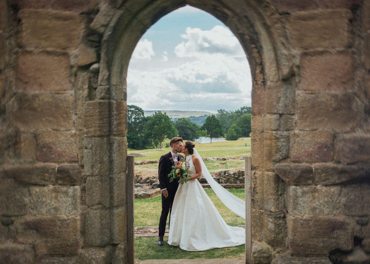 Bride and groom seen kissing through the arched stone doorway of Bolton Abbey